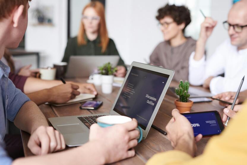 Besprechung von mehreren Personen an einem Tisch, Laptop mit Pr&auml;sentation und Notizen sichtbar.