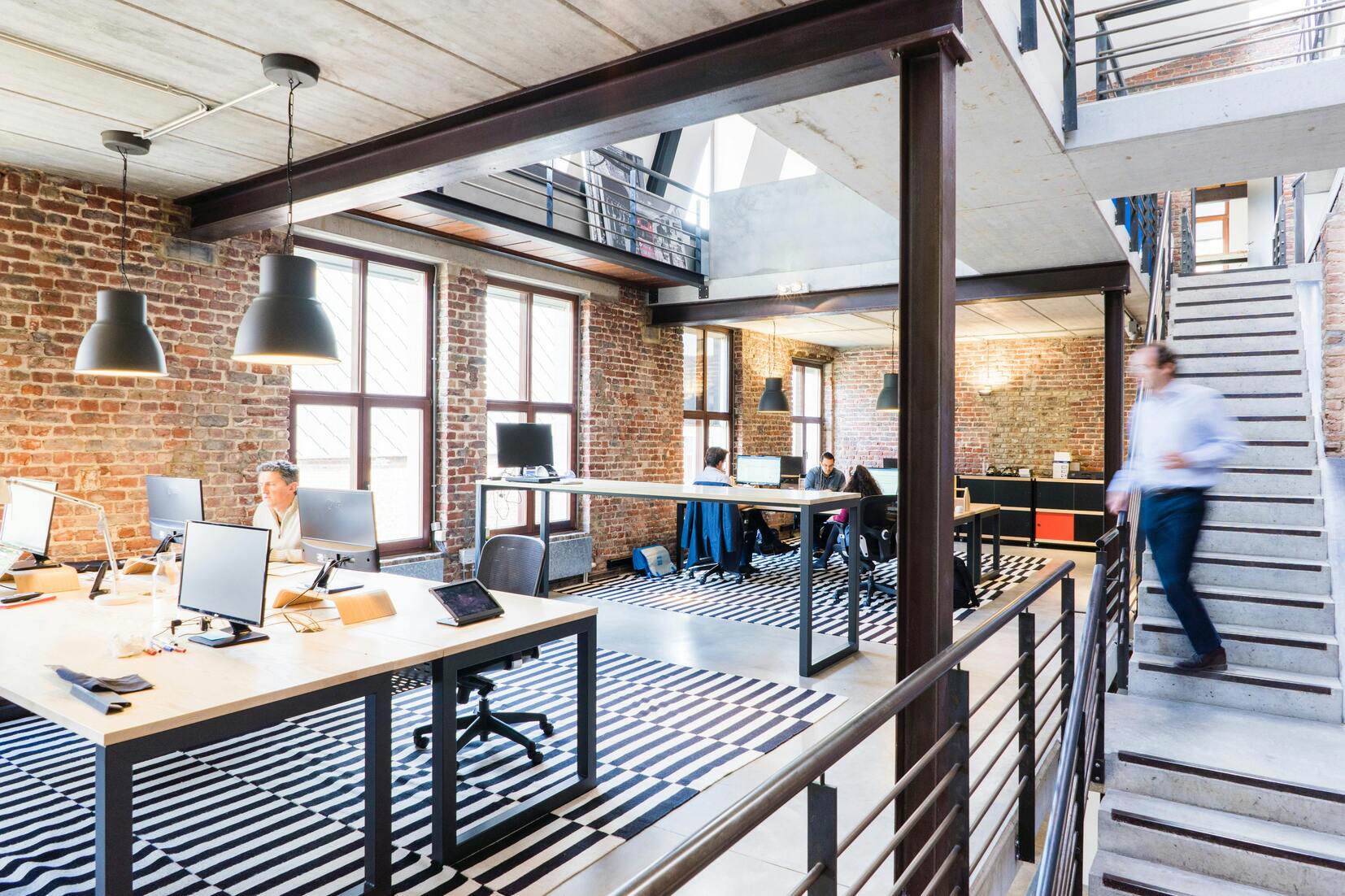 Offenes B&uuml;ro mit Holz- und Metallm&ouml;beln, gro&szlig;en Fenstern und einem zentralen Treppenhaus. Personen arbeiten am Computer.