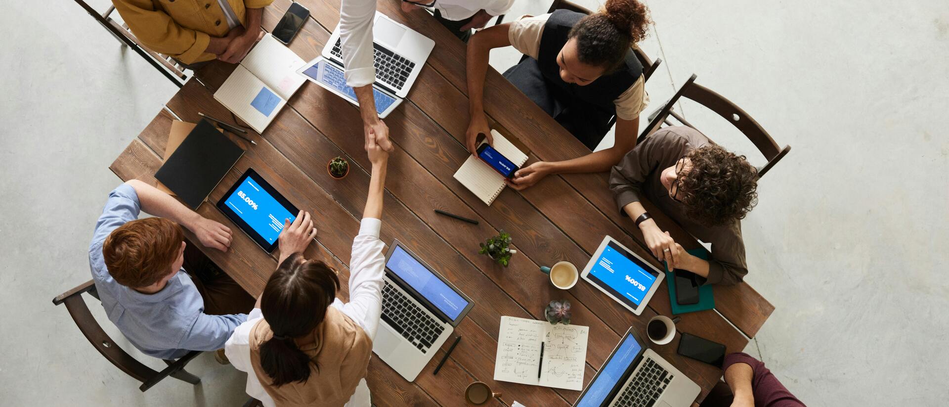 Vogelperspektive auf ein Meeting mit mehreren Personen, Laptops und Smartphone, die sich die Hand geben.