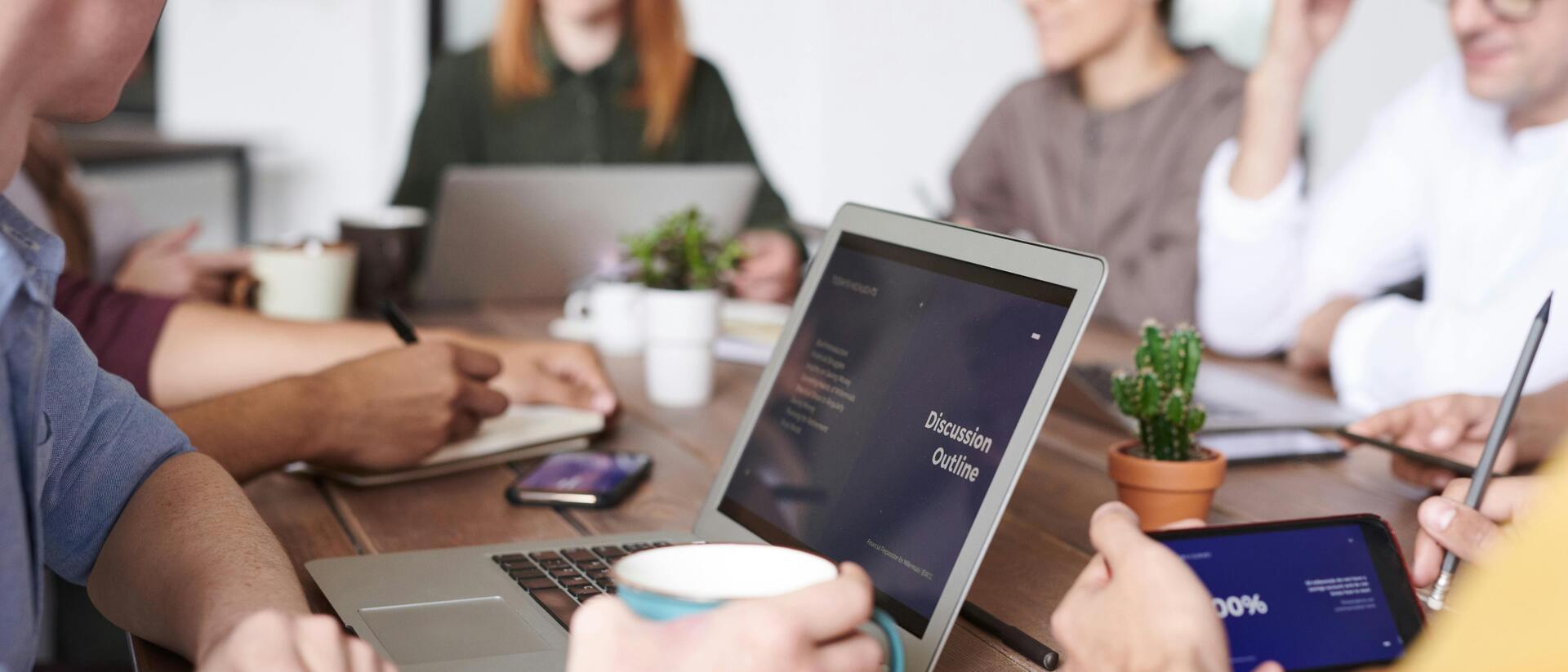 Besprechung von mehreren Personen an einem Tisch, Laptop mit Pr&auml;sentation und Notizen sichtbar.