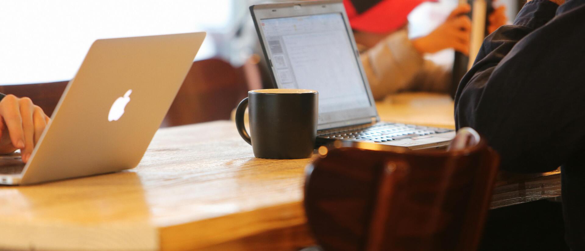 Zwei Laptops und eine Kaffeetasse stehen auf einem Holztisch, während Personen im Hintergrund beschäftigt sind.
