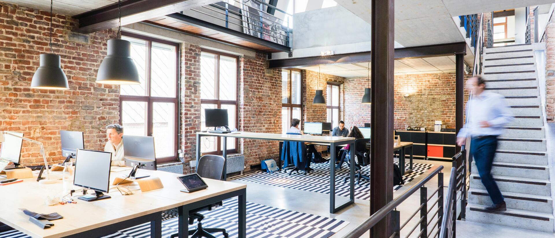 Offenes Büro mit Holz- und Metallmöbeln, großen Fenstern und einem zentralen Treppenhaus. Personen arbeiten am Computer.
