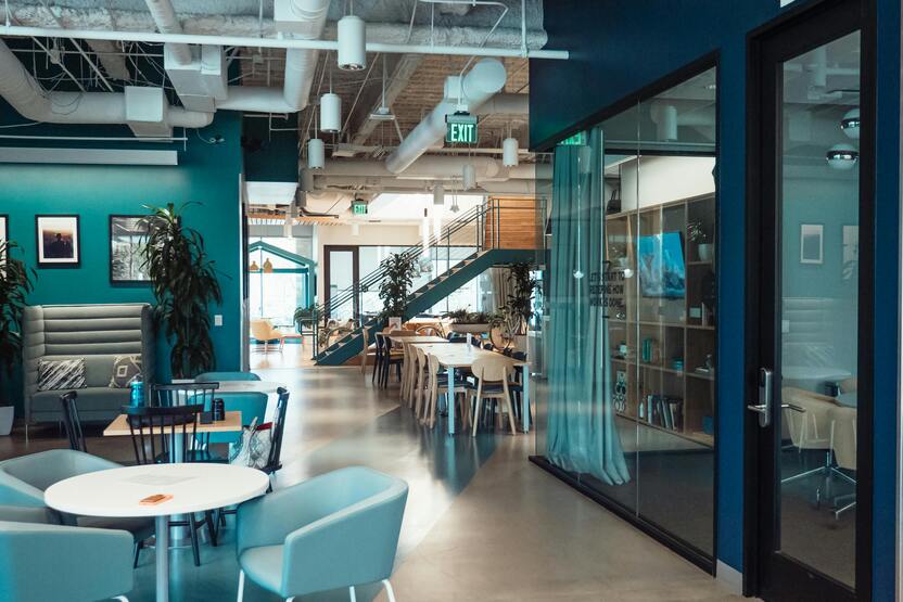Modernes B&uuml;ro mit blauen W&auml;nden, Tischen, St&uuml;hlen und einer Treppe im Hintergrund, gro&szlig;e Fenster und Pflanzen sichtbar.