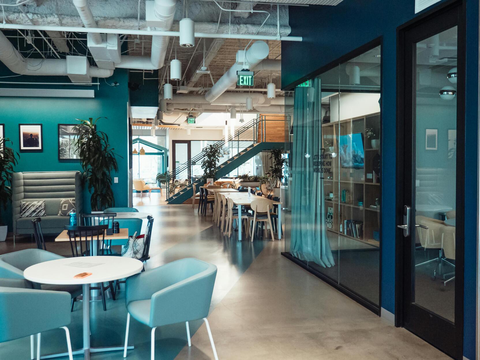 Modernes Büro mit blauen Wänden, Tischen, Stühlen und einer Treppe im Hintergrund, große Fenster und Pflanzen sichtbar.