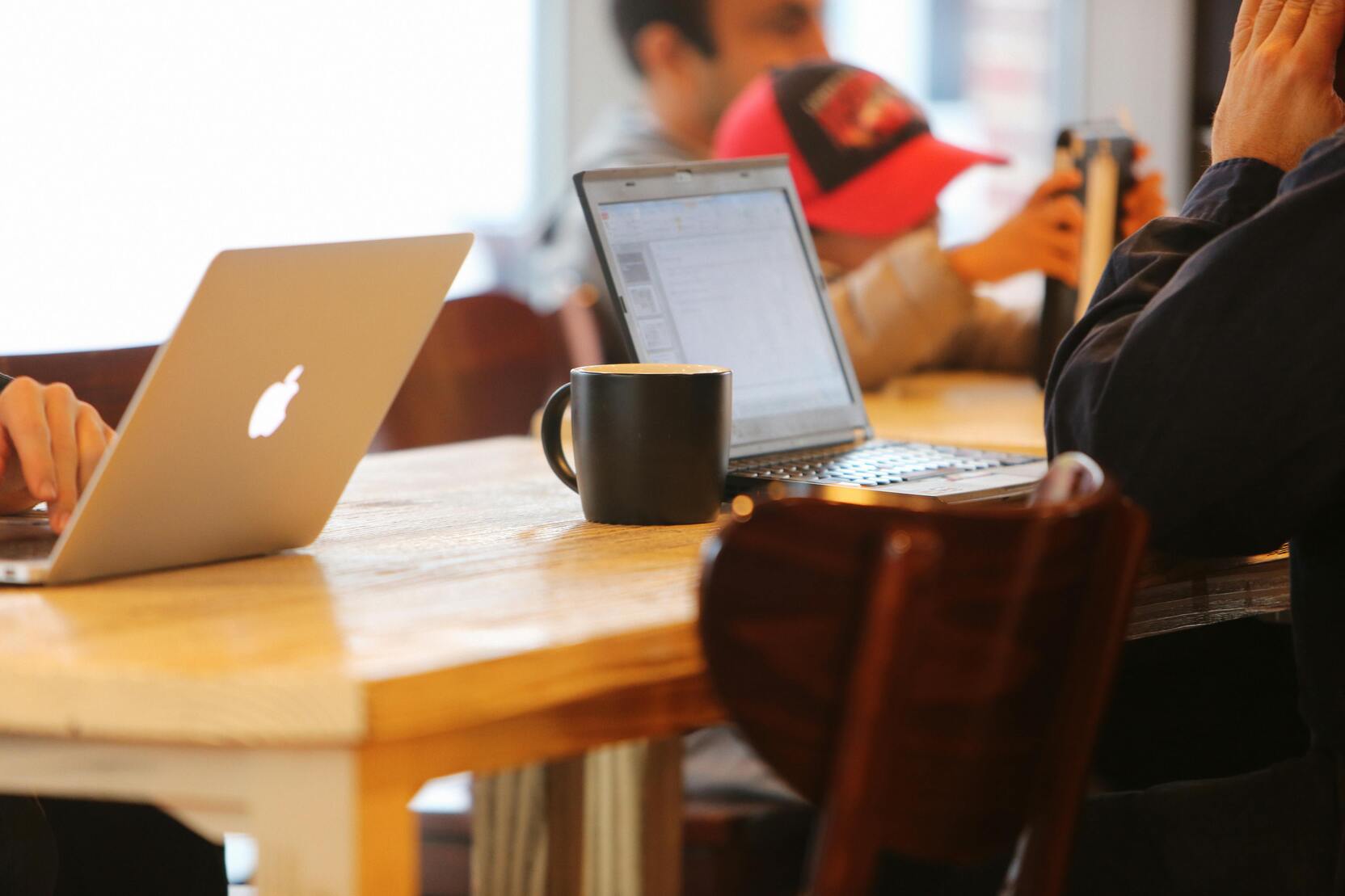 Zwei Laptops und eine Kaffeetasse stehen auf einem Holztisch, während Personen im Hintergrund beschäftigt sind.
