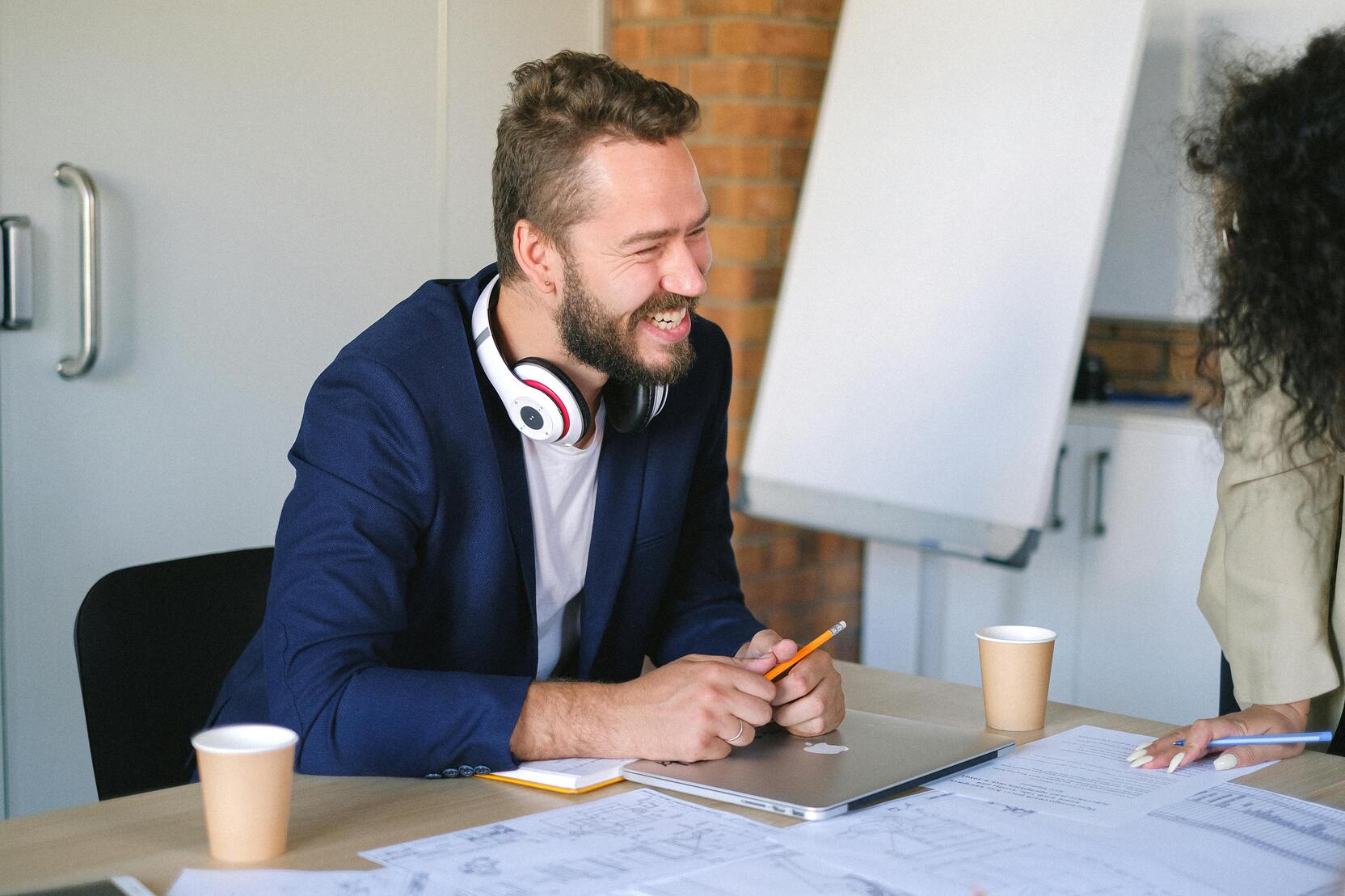 Lachender Mann mit Kopfh&ouml;rern sitzt an einem Tisch, umgeben von Kaffeebechern und Dokumenten.